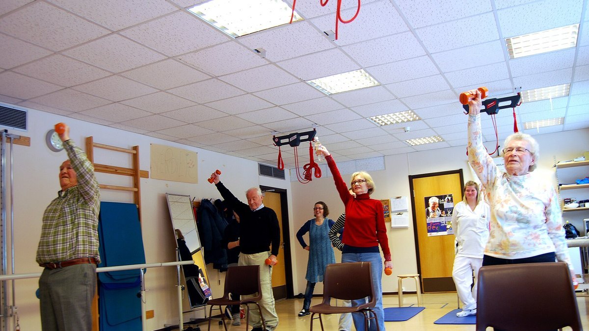 Older men and women in an exercise room lift light hand weights overhead, with chairs in front of them for support