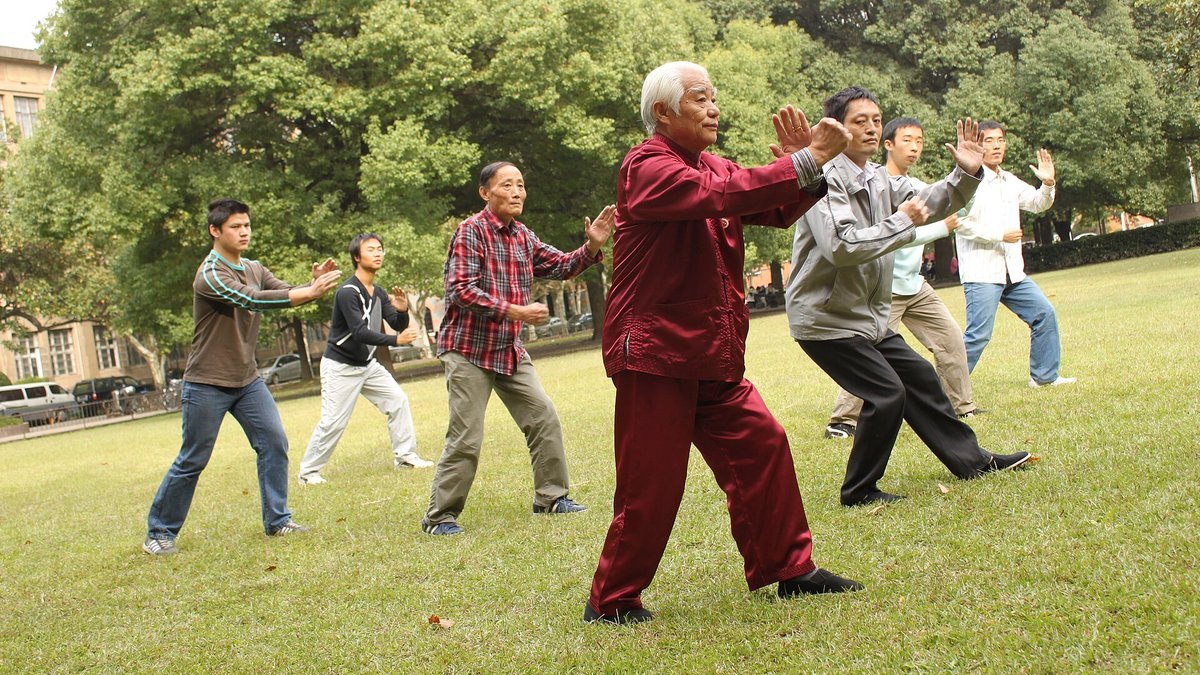 An older man in a red tai chi outfit leads a group of people of mixed ages through a slow tai chi posture on a park lawn