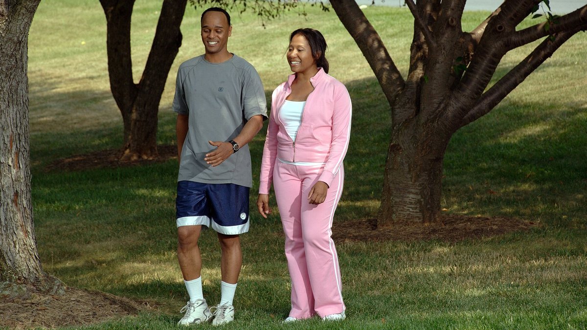 A smiling man in a gray T-shirt and navy shorts and a woman in a pink tracksuit walk together across the grass in a sunny, tree-shaded park