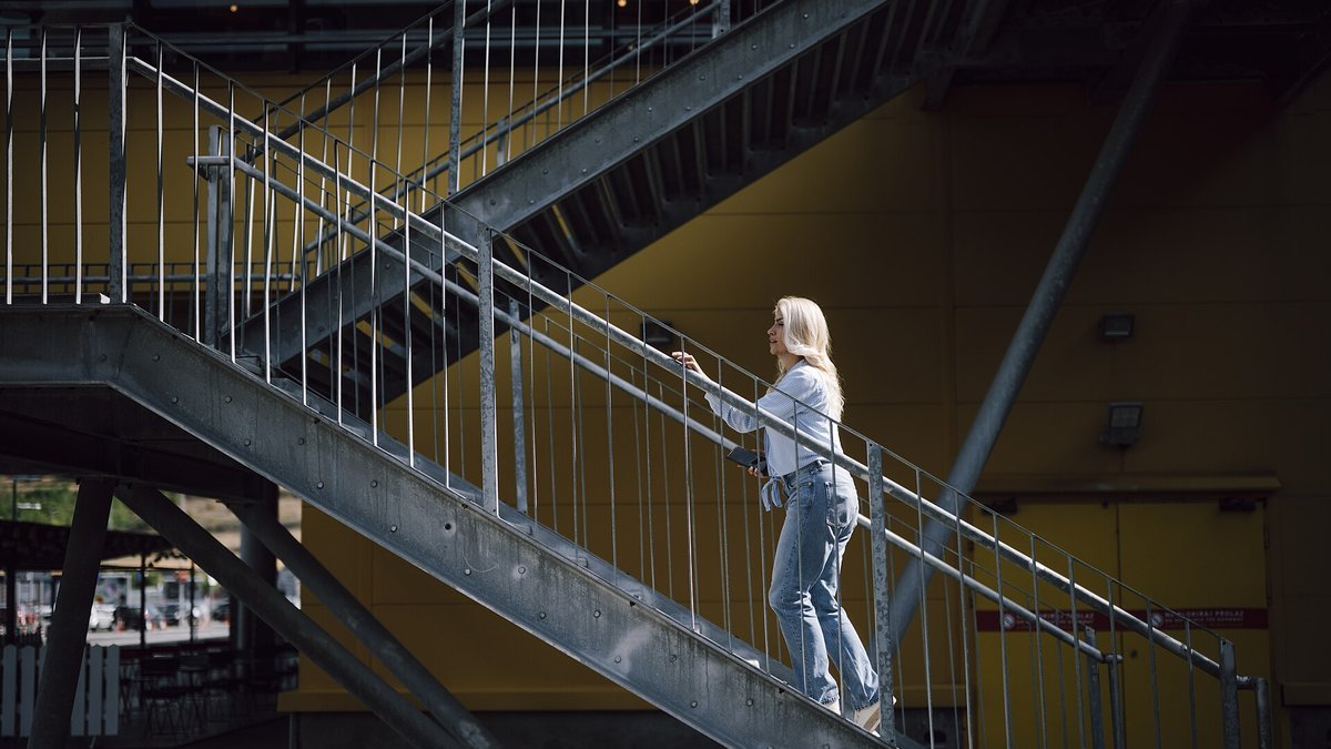 A woman in jeans walks up an outdoor metal staircase alongside a yellow building, holding the handrail