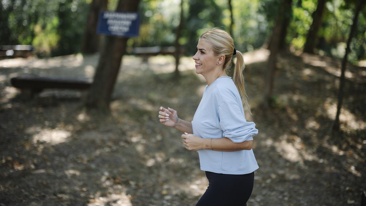 A smiling woman with a blonde ponytail, in a light blue sweatshirt and black leggings, jogs along a shaded park path with trees and benches behind her