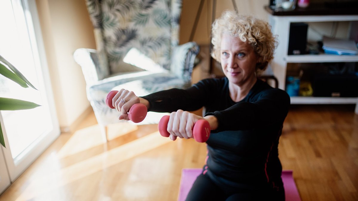 A woman with curly blond hair sits on a pink exercise mat in a sunlit living room, holding a pair of light pink dumbbells straight out in front of her