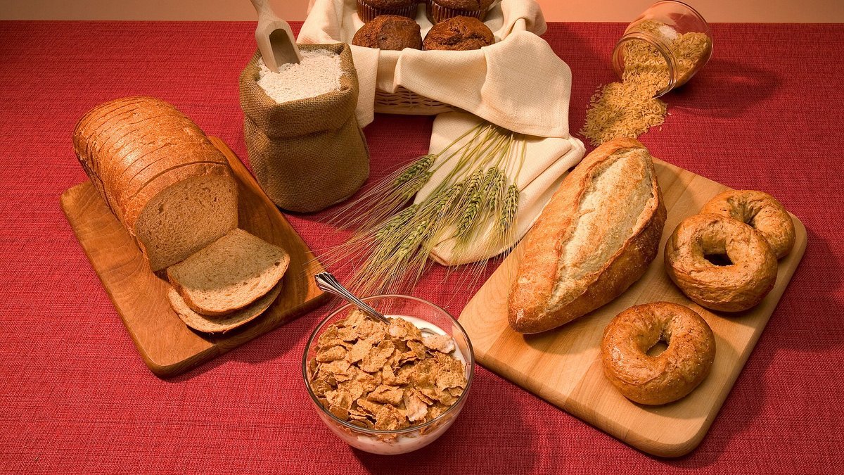 Whole-grain foods on a red tablecloth: a sliced whole-wheat loaf, a bowl of whole-grain flake cereal, whole-wheat bagels, bran muffins, a crusty loaf, wheat stalks, a sack of whole-wheat flour and a jar of brown rice