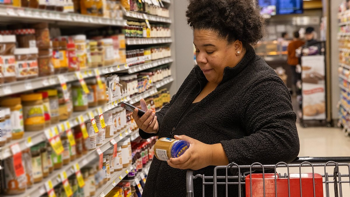 A shopper in a grocery aisle lined with jars and packaged foods holds a jar in one hand and checks product information on her phone with the other