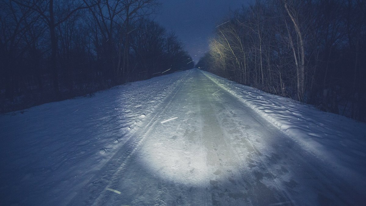 A headlamp beam lights up a snow-covered, icy trail between bare trees at night while snow falls