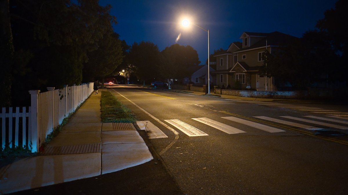 A quiet residential street at night with a streetlight, a sidewalk beside a white picket fence, a curb ramp and a marked crosswalk