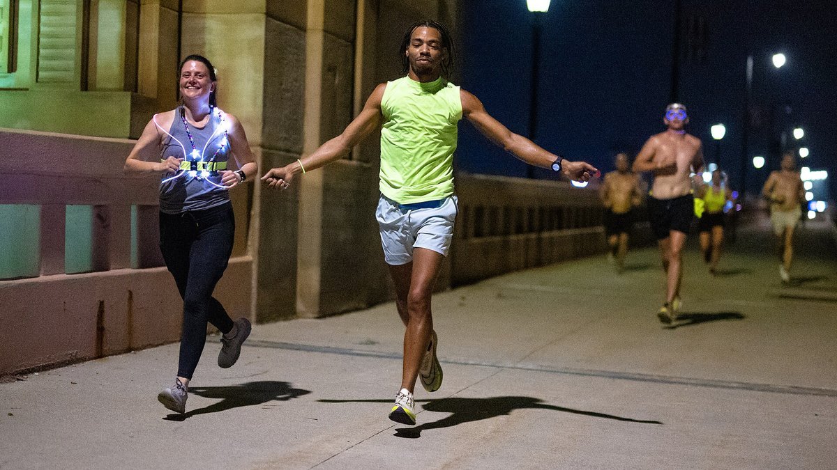 Two runners, one in a neon yellow tank top and one wearing a light-up harness, run along a lit bridge walkway at night, with more runners and streetlights behind them