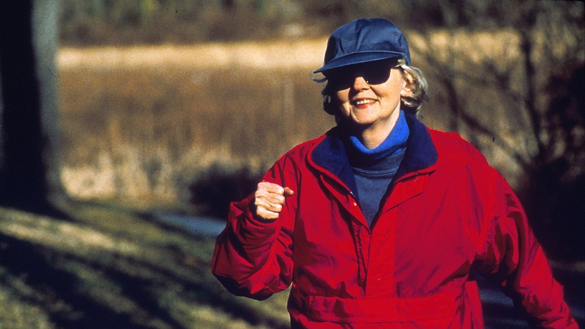 An older woman in a red jacket, blue cap and sunglasses smiles as she power walks along a sunny wooded path, arms bent and swinging