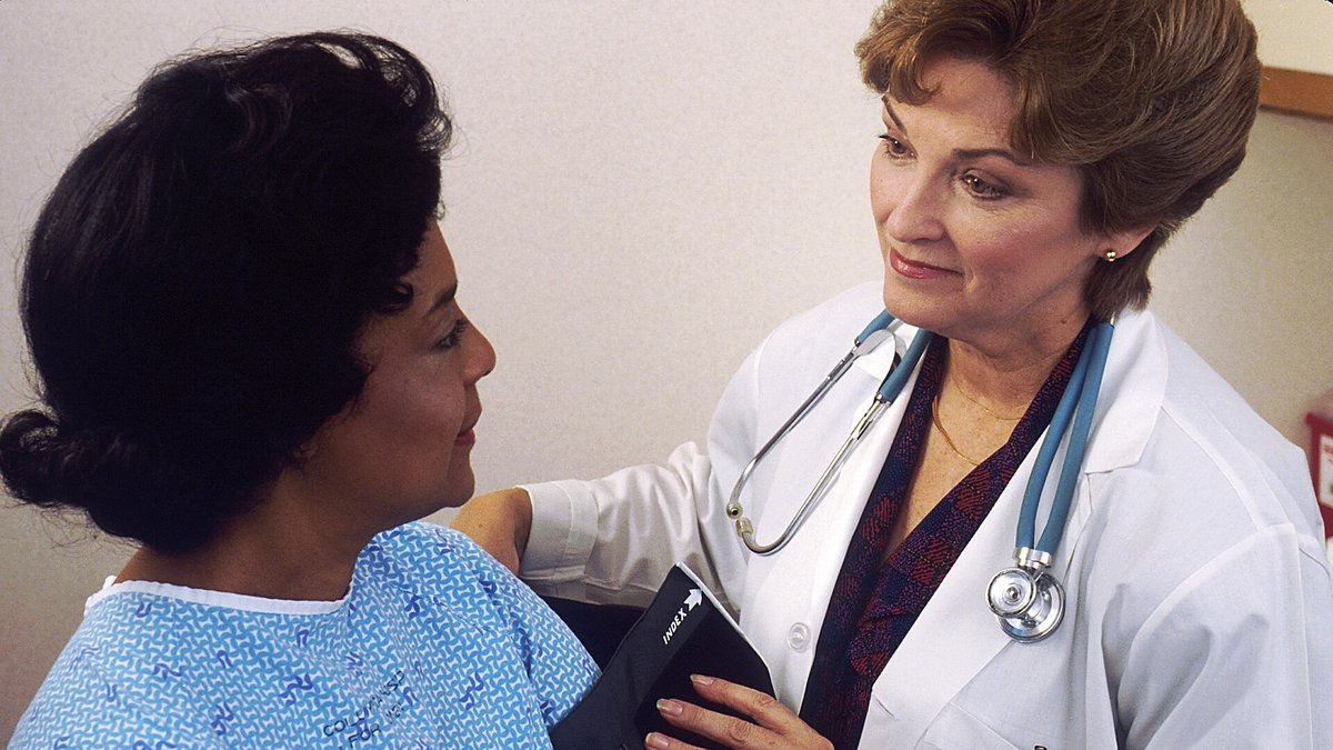 A doctor in a white coat with a stethoscope places a blood pressure cuff on the upper arm of a seated woman in a blue patient gown