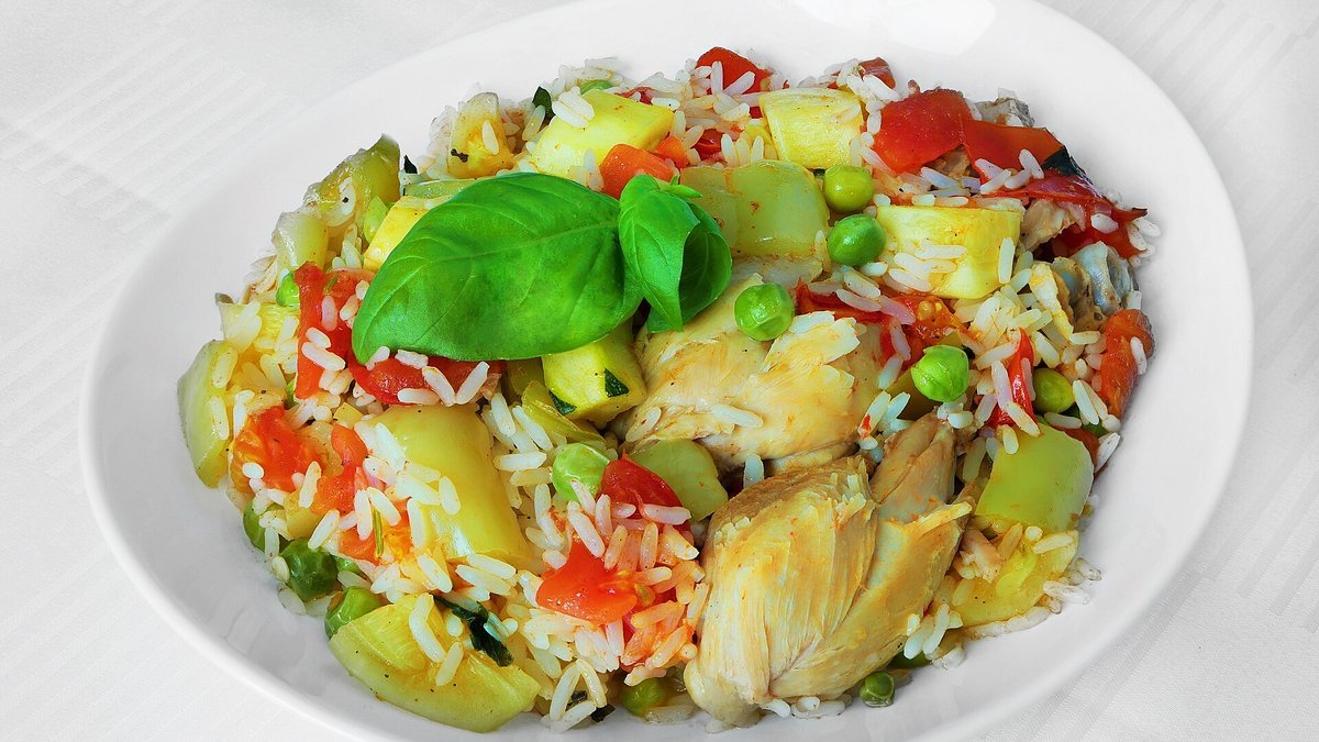 A white bowl of rice mixed with chicken, peppers, squash, peas and tomato, topped with basil leaves.