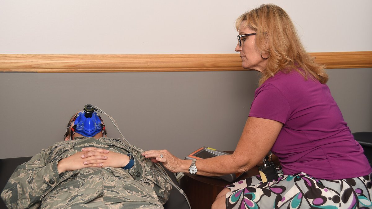 A health program coordinator sits beside an airman lying on her back with a mask over her face during a resting metabolic rate test.